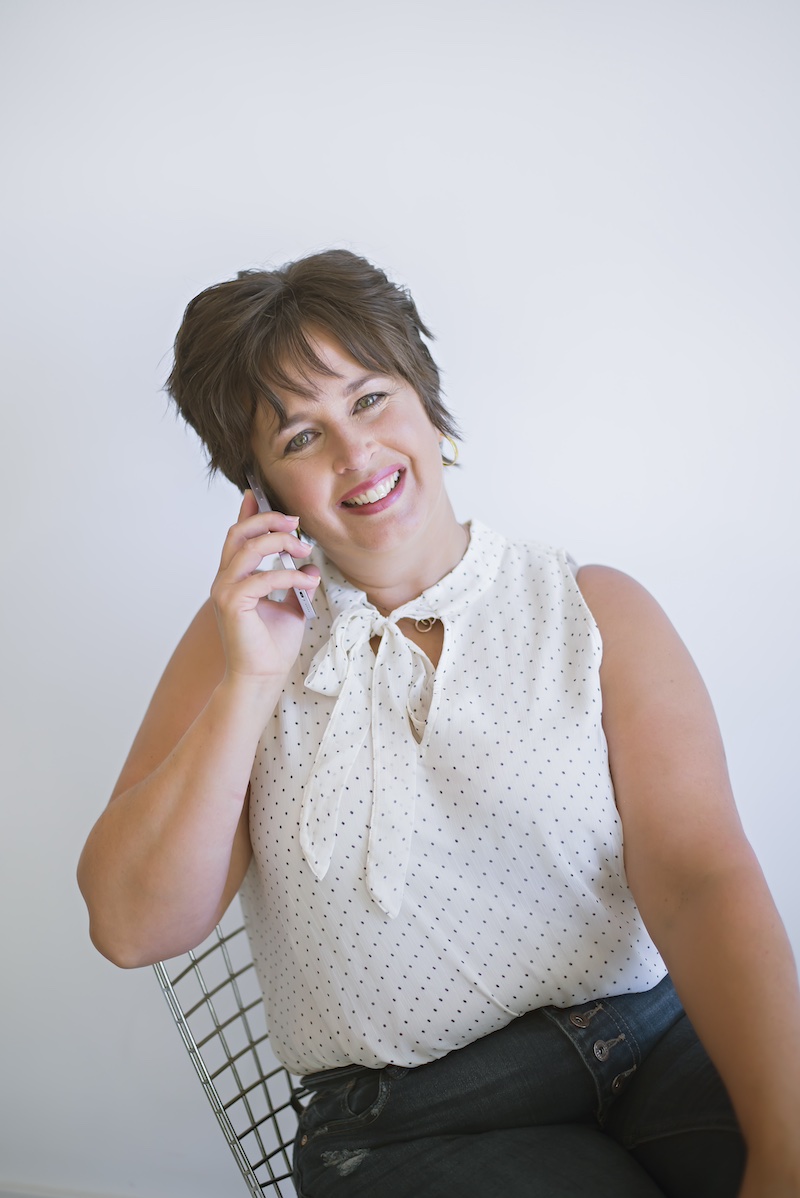 Kim Hagle, body positive personal trainer in Goderich, ON, smiling while talking on the phone, wearing a white polka-dot blouse and seated on a wire chair against a plain white background